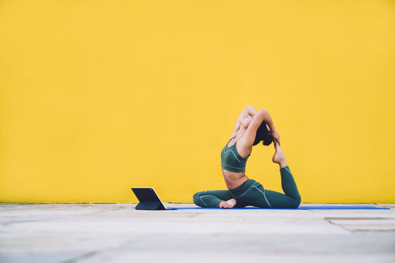 Side view of attractive flexible healthy female in sports outfit sits in one legged king pigeon posture on yoga mat near tablet on yellow background